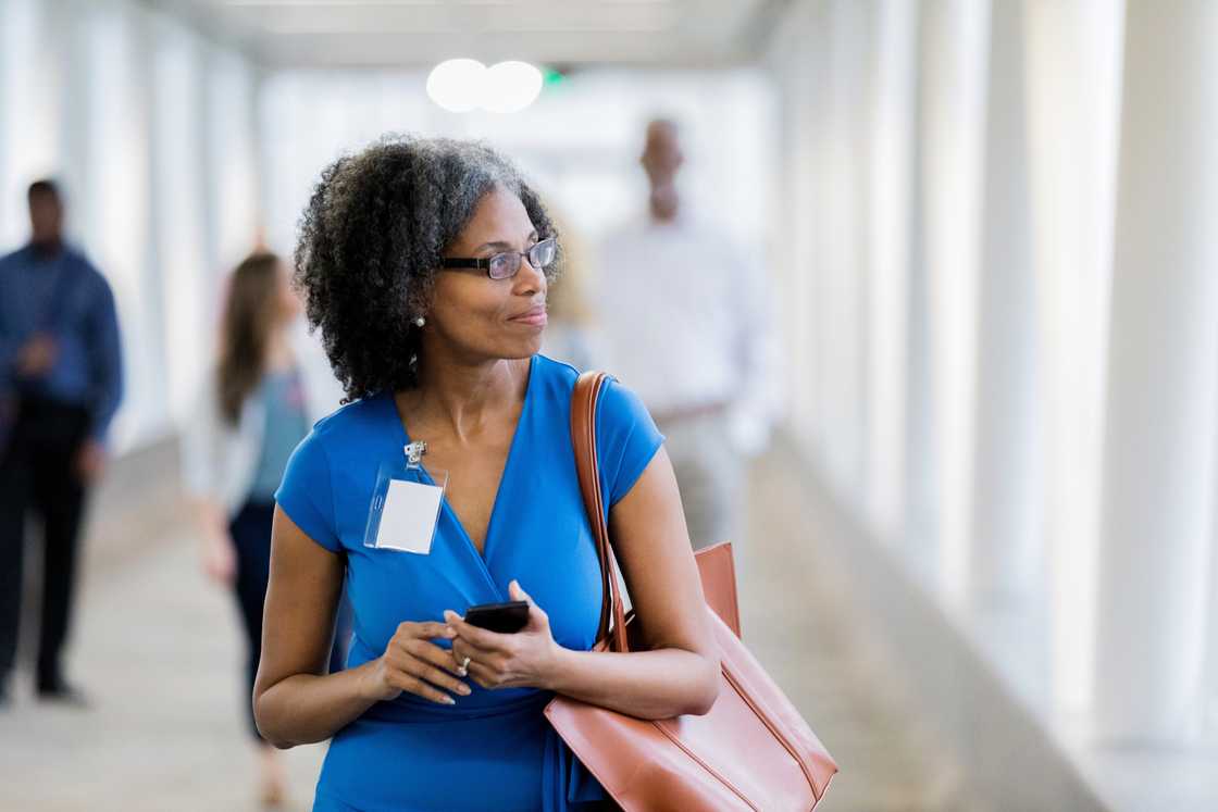 Woman in a blue dress stands in a bright corridor holding a phone and tote bag.