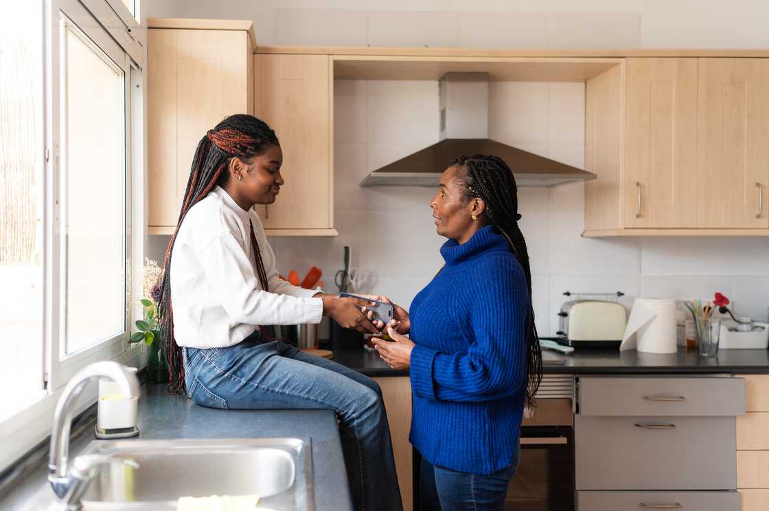Two people talk in a sunlit kitchen, one seated on the counter holding a phone, the other standing and listening.