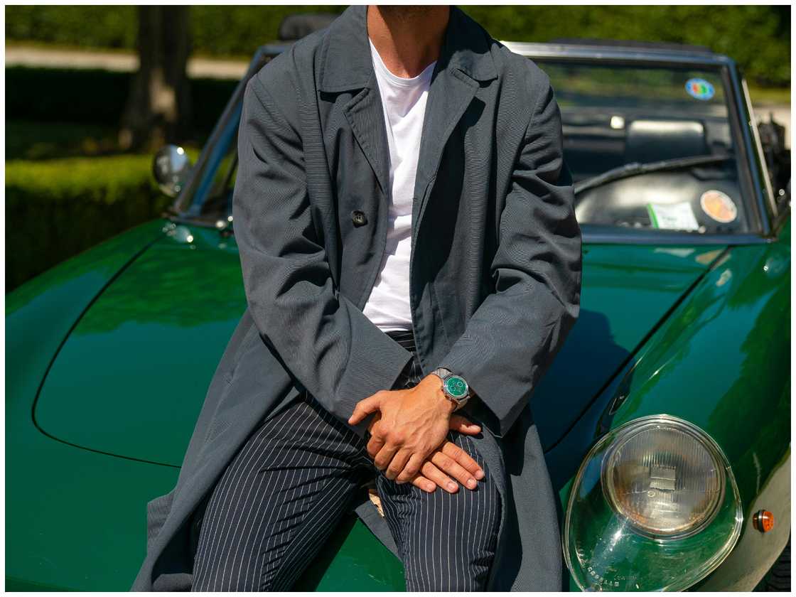 A man in a grey trench coat leaning on a green car