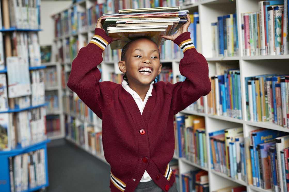 A schoolgirl smiling and balancing a stack of books on her head in a library