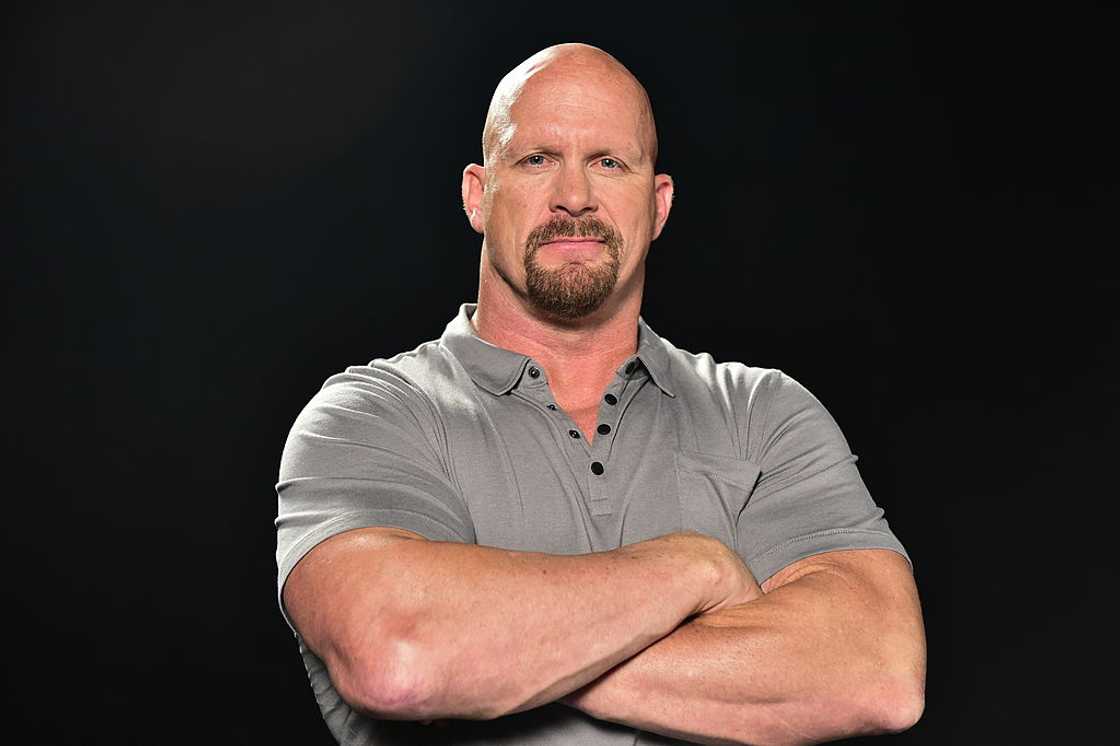 Steve Austin Stone Cold, one of the 90s wrestlers, is wearing a grey t-shirt and folding his hands as he poses for a photo.