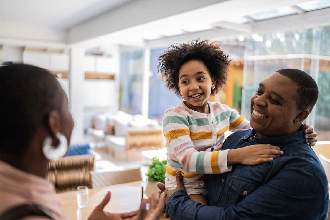 Smiling man holds a young girl in pastel stripes as they greet a woman indoors near large windows. Smiling man holds a young girl in pastel stripes as they greet a woman indoors near large windows.
