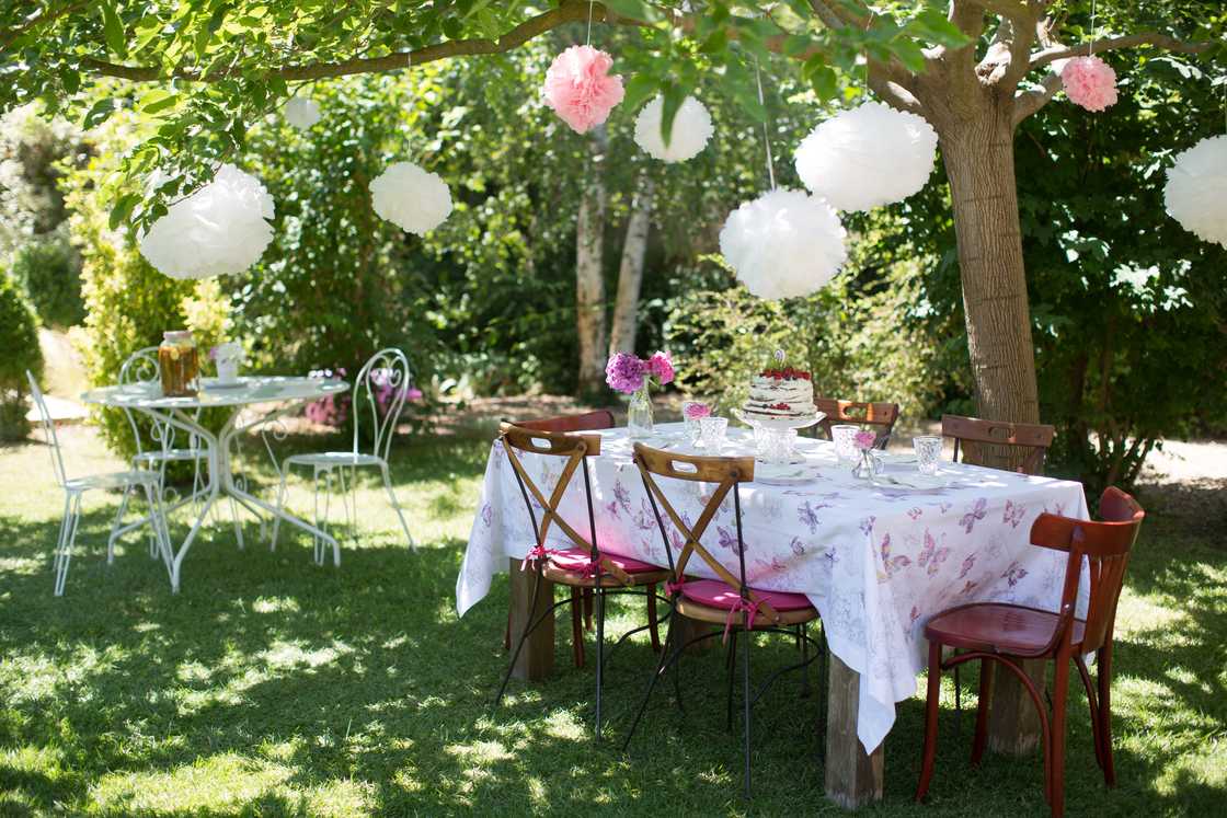 Outdoor table set for a garden party with pink floral decor, hanging lanterns, and a layered cake centrepiece. Outdoor table set for a garden party with pink floral decor, hanging lanterns, and a layered cake centrepiece.