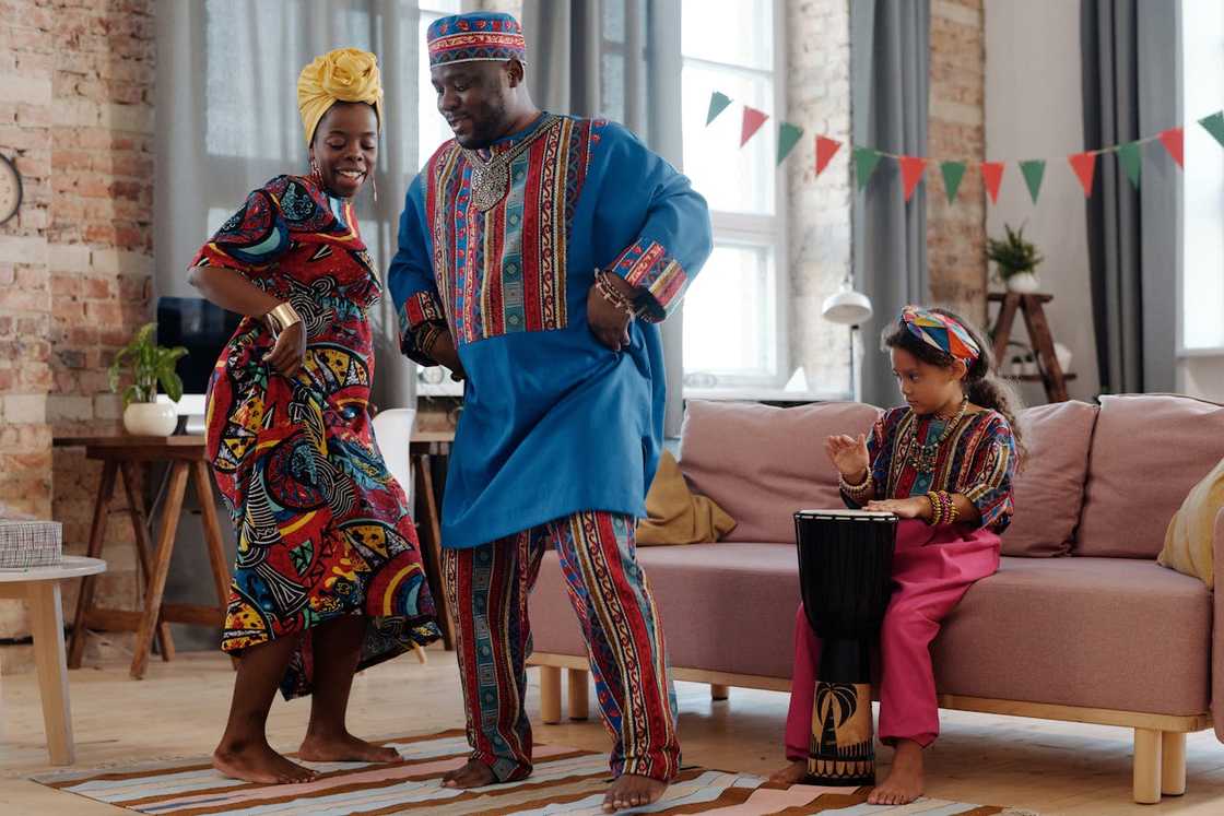 A family in colourful traditional clothing dances in a living room while a child plays a drum.