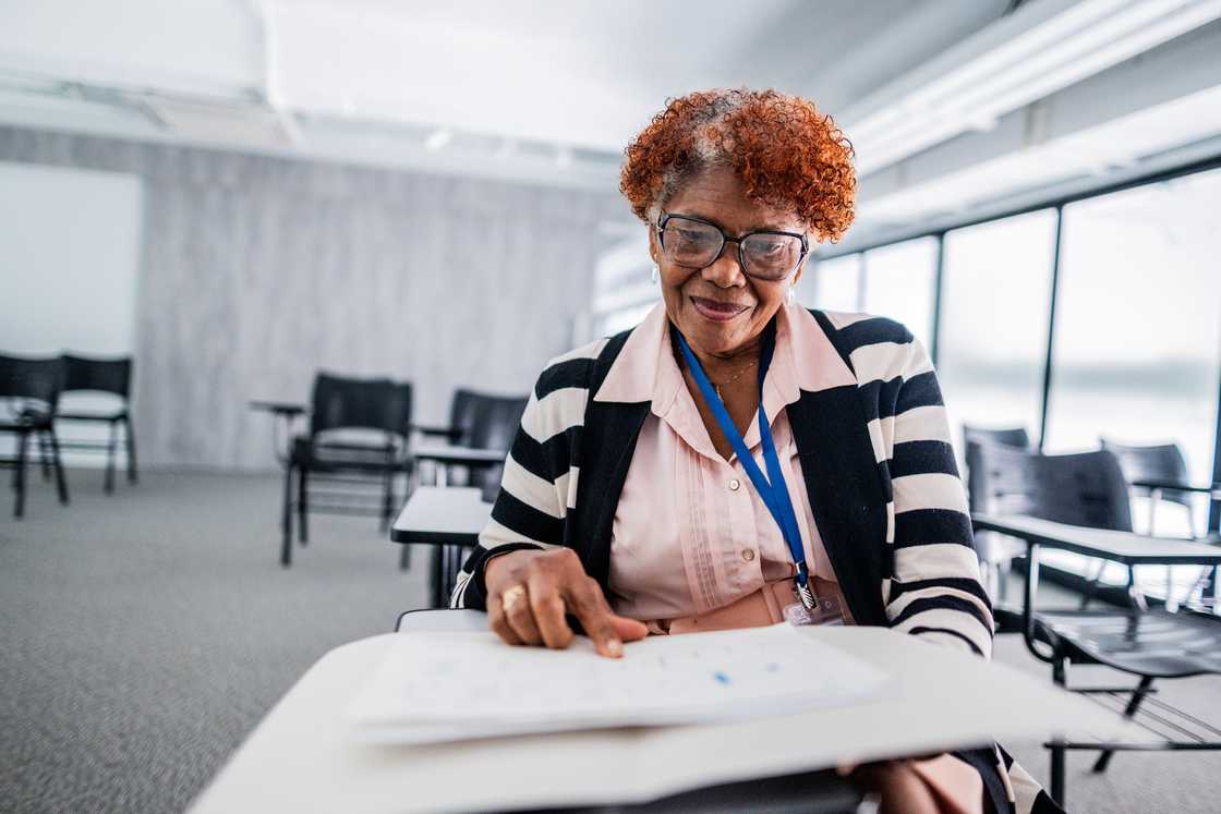Older adult in glasses points at a worksheet while seated in a bright classroom. Older adult in glasses points at a worksheet while seated in a bright classroom.