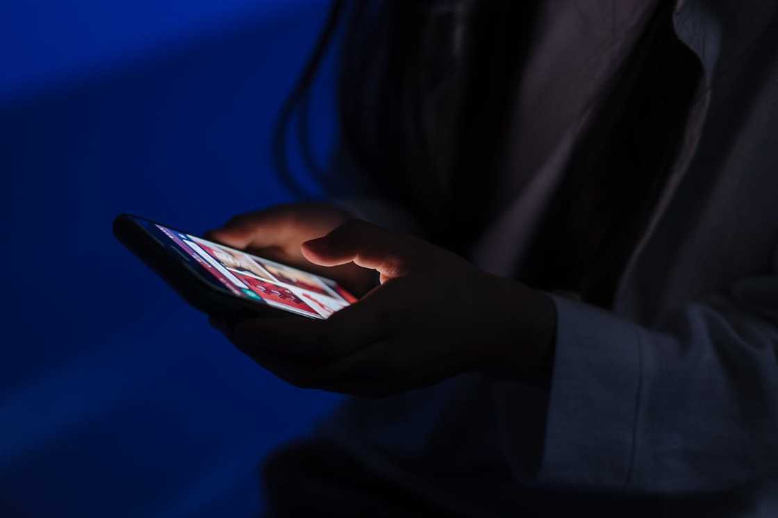A person holds a phone in a dark room, the screen glowing in the blue light. A person holds a phone in a dark room, the screen glowing in the blue light.