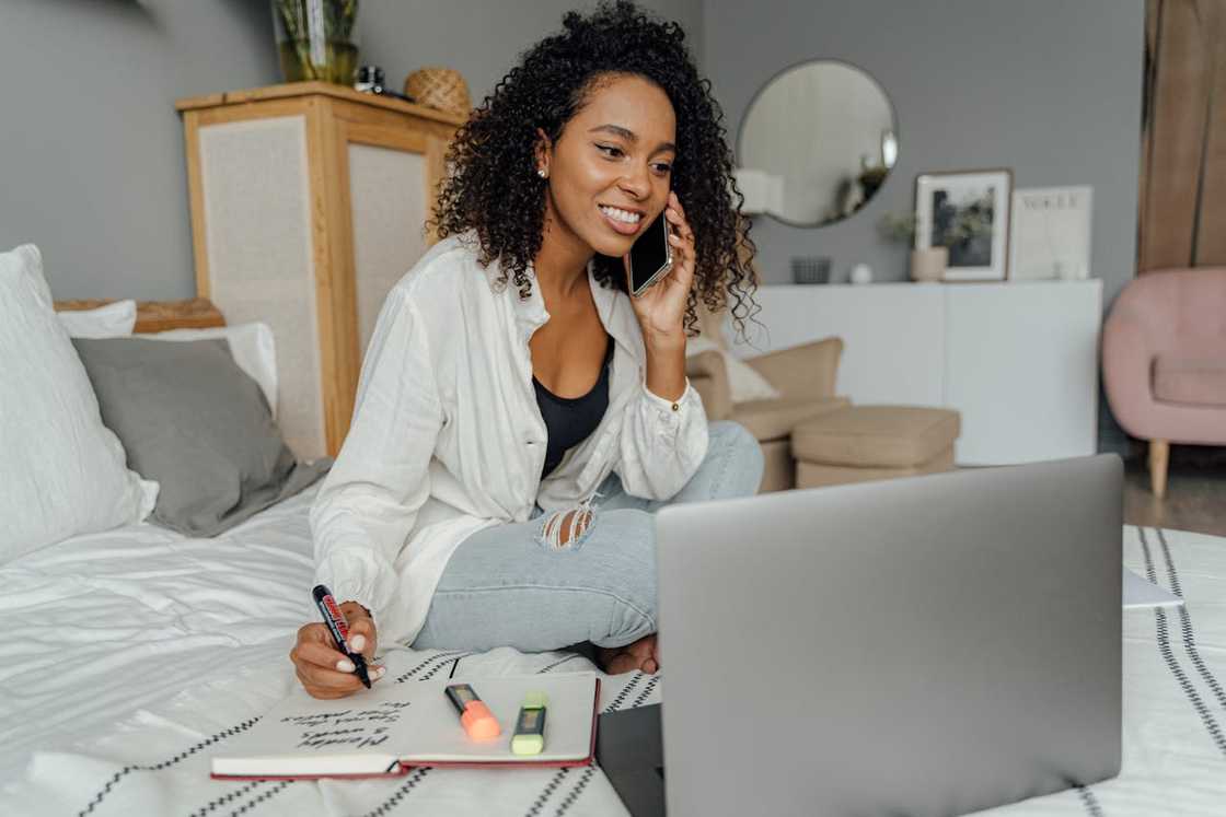 A woman smiling while talking on the phone and working on a laptop at home.