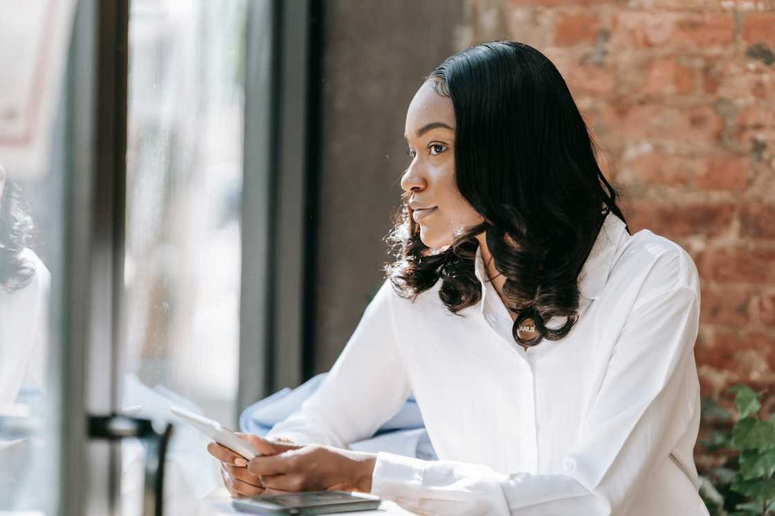 A woman sits by a window holding a phone and looking outside thoughtfully.