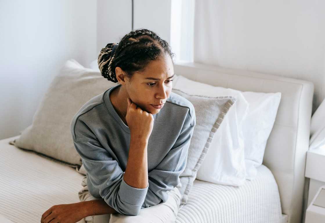 A woman sits on a bed, resting her chin on her hand, looking thoughtful in a softly lit bedroom.