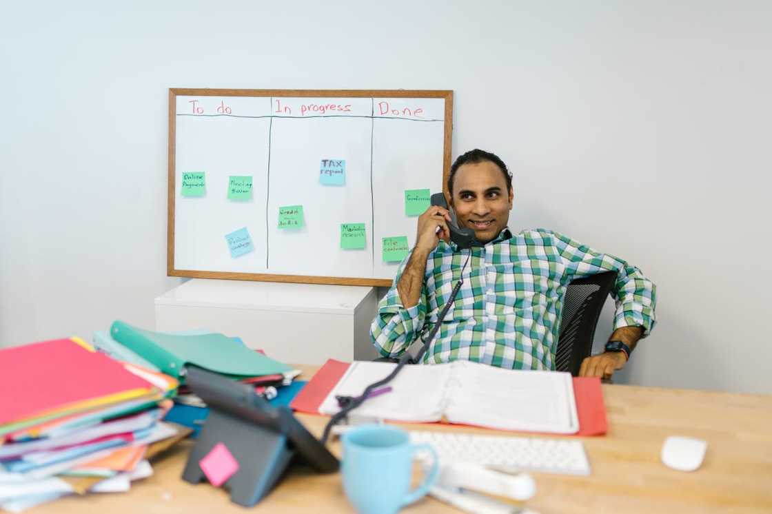 A man multitasks on the phone, surrounded by documents and a task board.