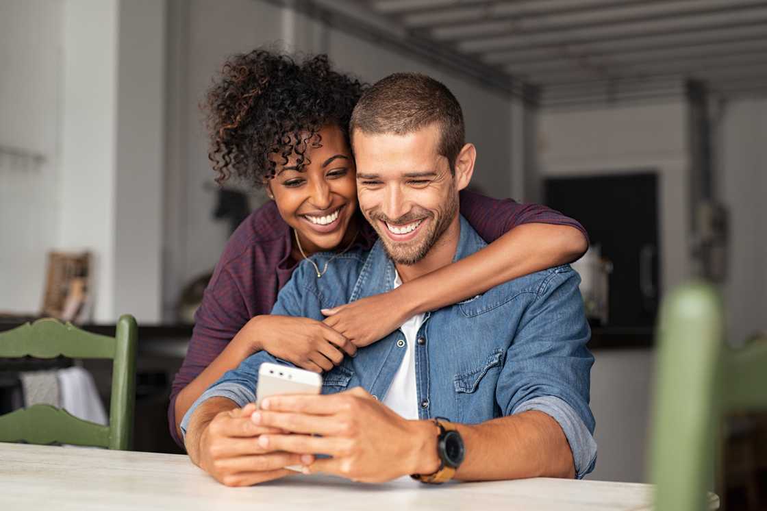 A smiling young couple embrace while looking at a smartphone.