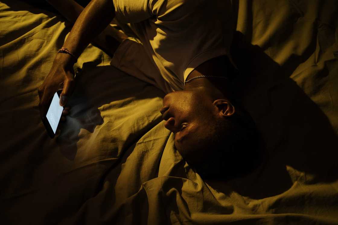 A person lying on a bed at night, holding a glowing phone in dim light.
