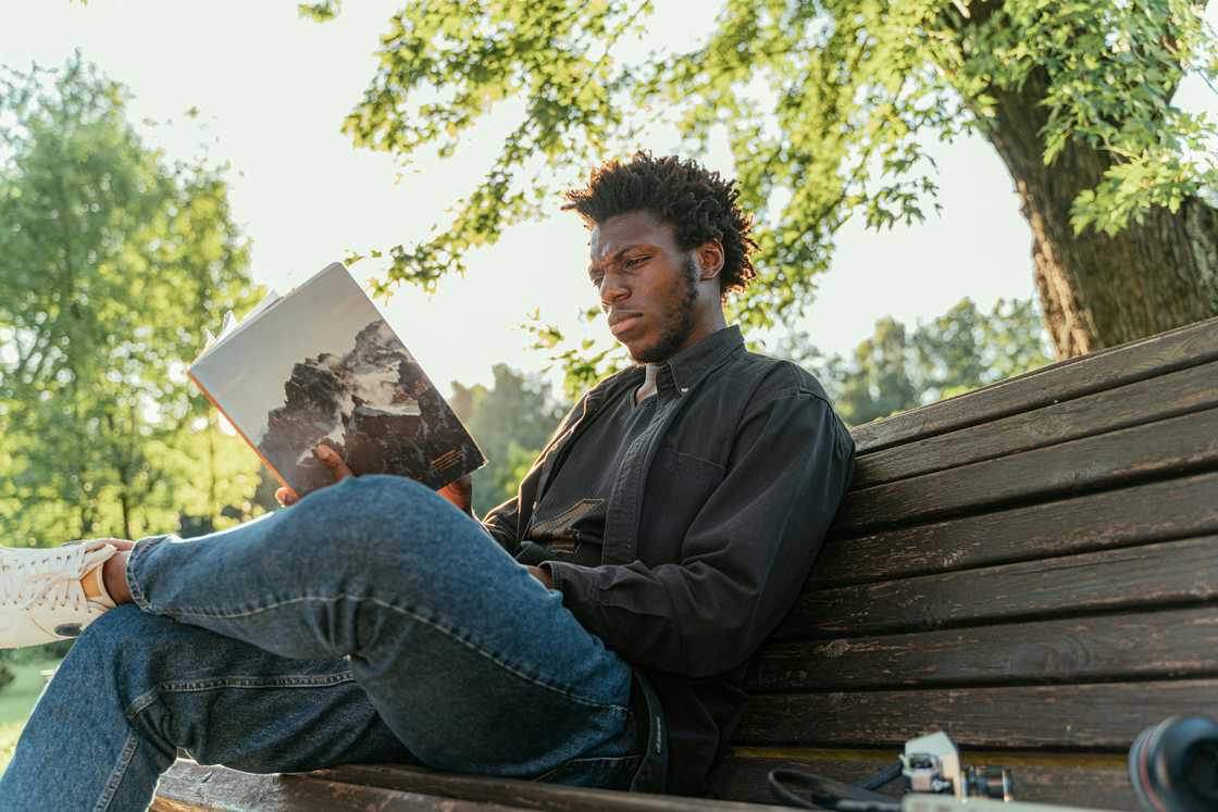 A young man in denim pants and a black shirt is reading a book on a bench