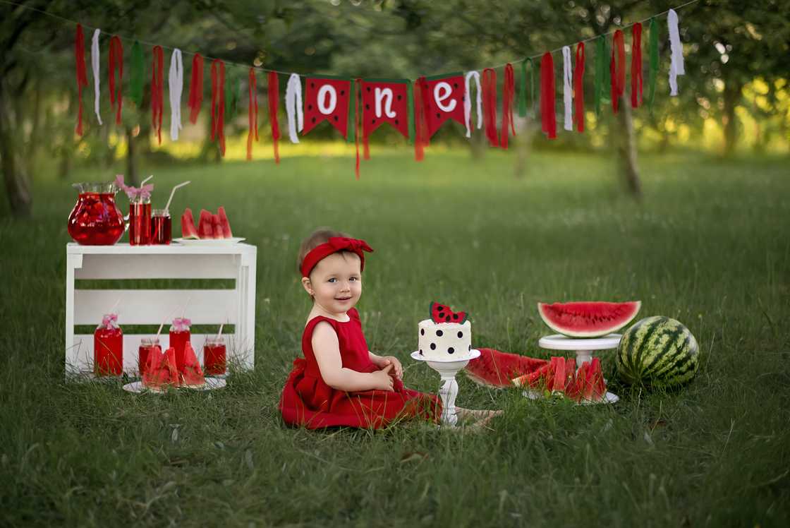 The baby girl celebrates her birthday. There is a cake and a lot of watermelons in front of the girl