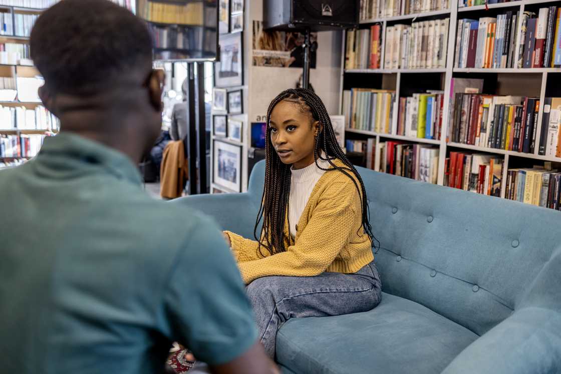 Two people talk on a couch in a room lined with bookshelves.