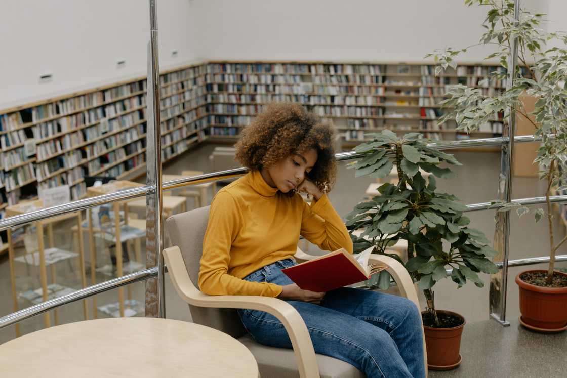 A woman in a yellow top and denim pants is reading a book in a library