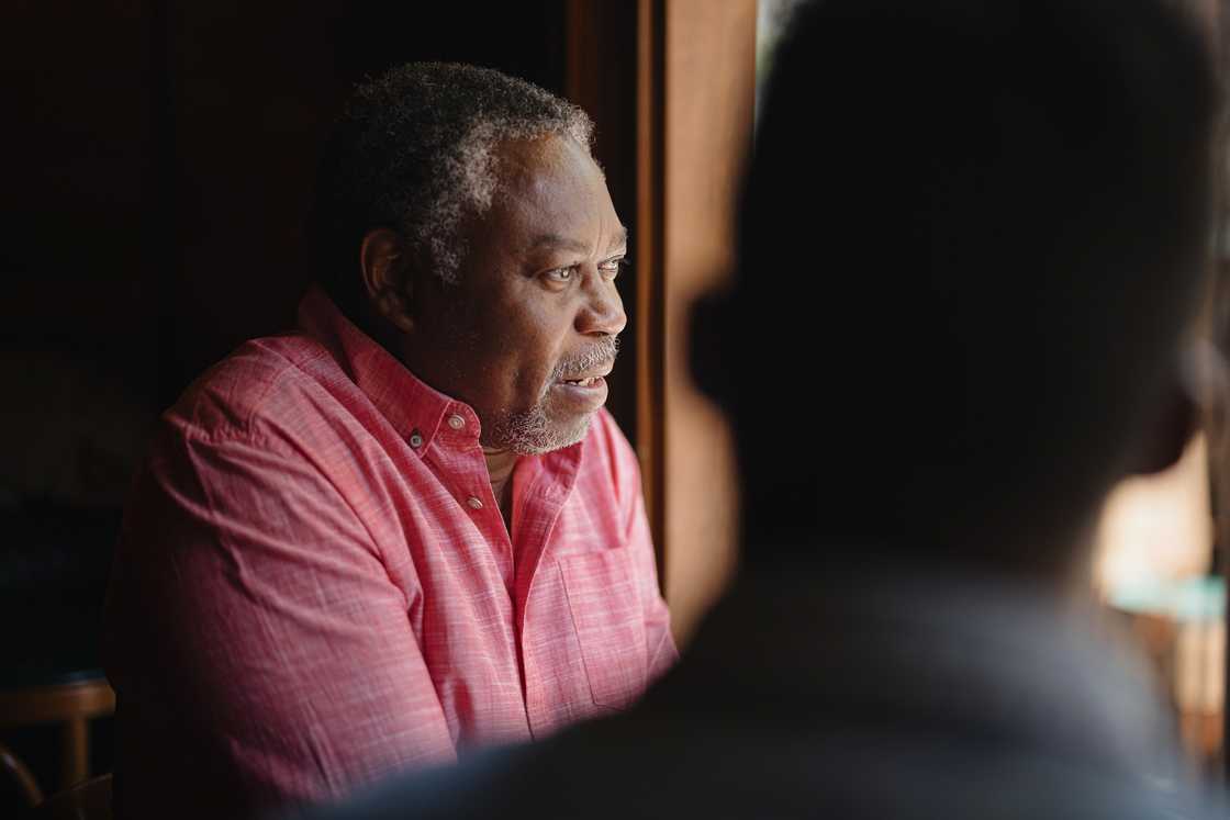An older man in a red shirt sits indoors near a window.