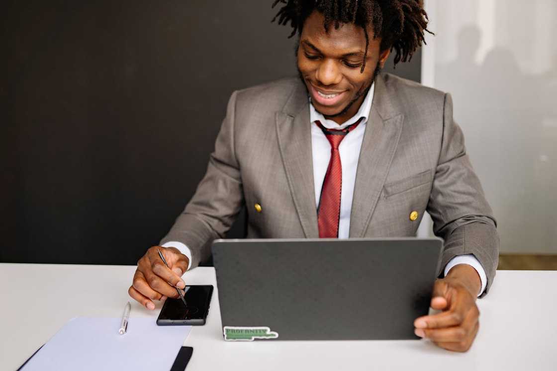 A man smiles while working on a laptop at a desk.