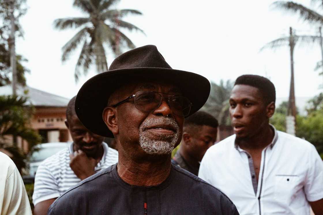 An older rich man wearing a hat standing outdoors with others behind him. An older rich man wearing a hat standing outdoors with others behind him.