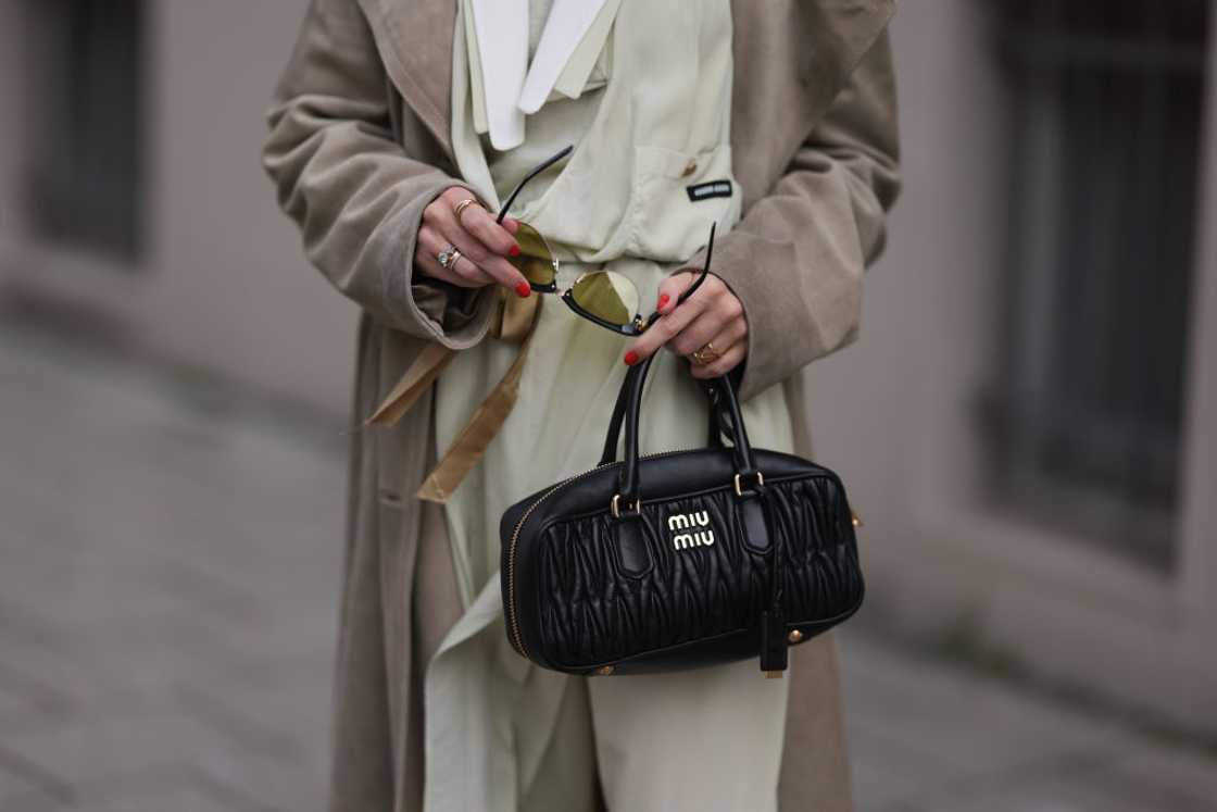 A stylish woman in a trench coat and sunglasses holding a black Miu Miu leather bag. A stylish woman in a trench coat and sunglasses holding a black Miu Miu leather bag.