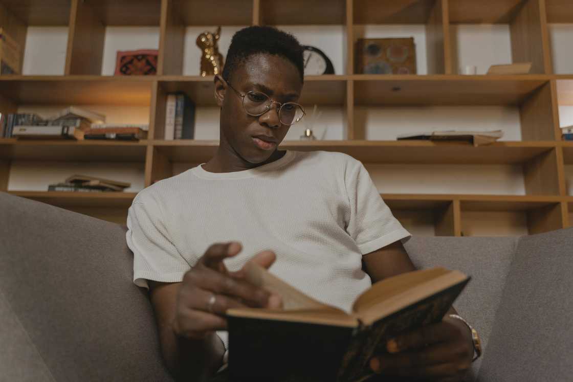 A young man in glasses is reading a book on the sofa