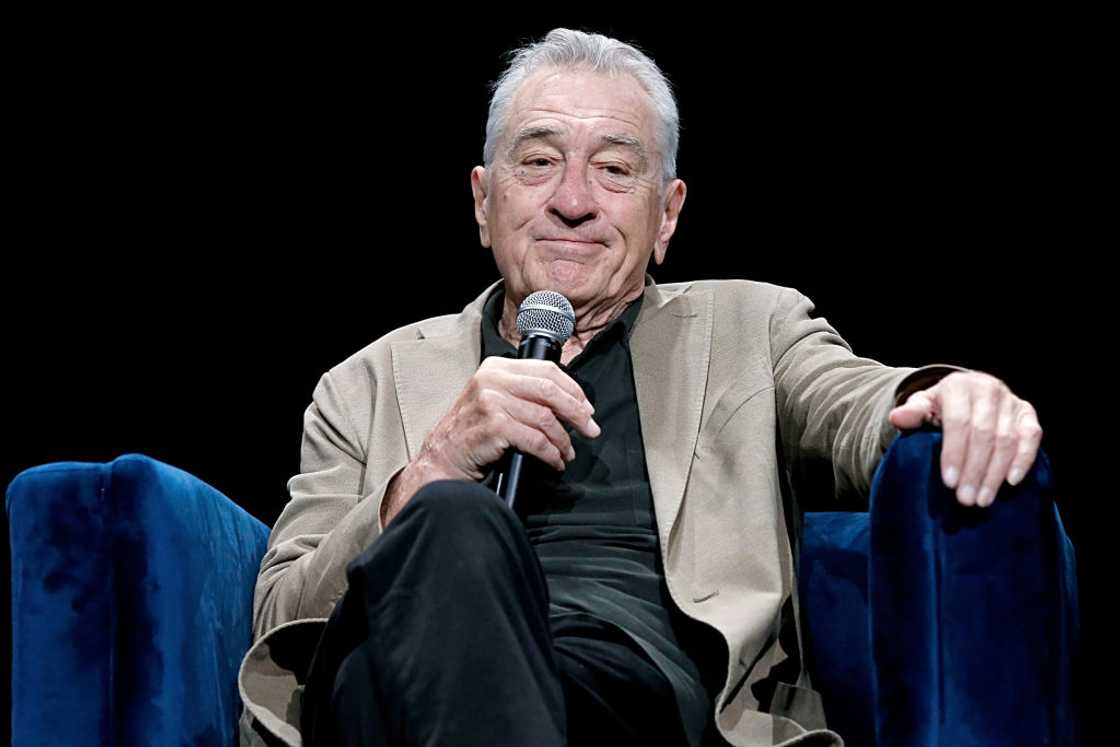 Robert De Niro in a grey coat and holding a microphone