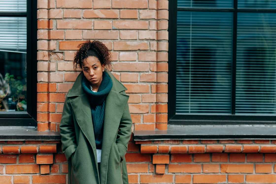 A woman stands against a brick wall between two windows.