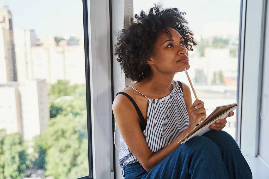 Person sits by a window holding a tablet and pencil, gazing upward in thought. Person sits by a window holding a tablet and pencil, gazing upward in thought.