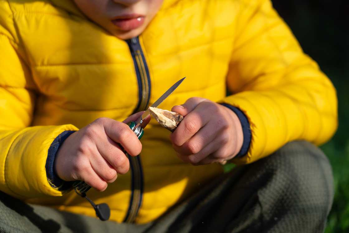 A boy sharps a stick with a utility knife to make an arrow. A boy sharps a stick with a utility knife to make an arrow.
