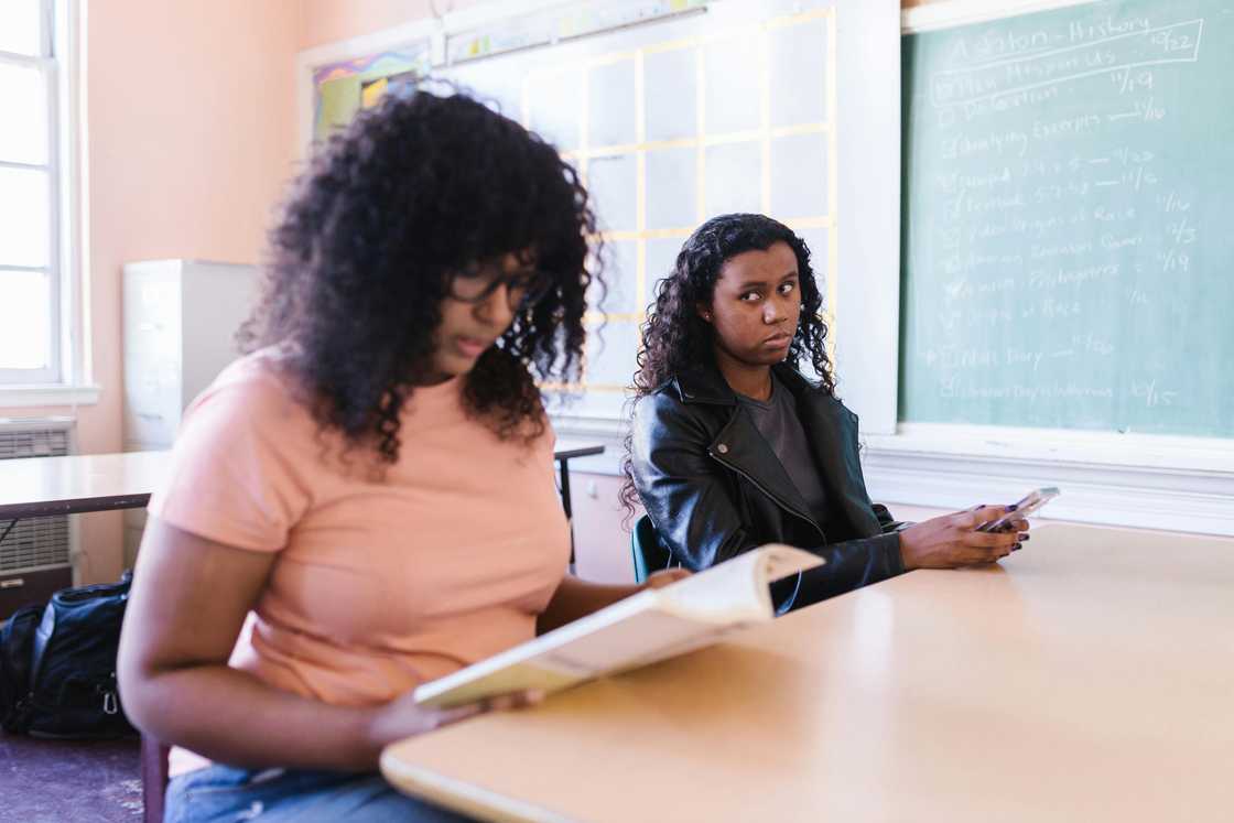 A woman in a black leather jacket is looking at another woman in a pink top as she reads a book