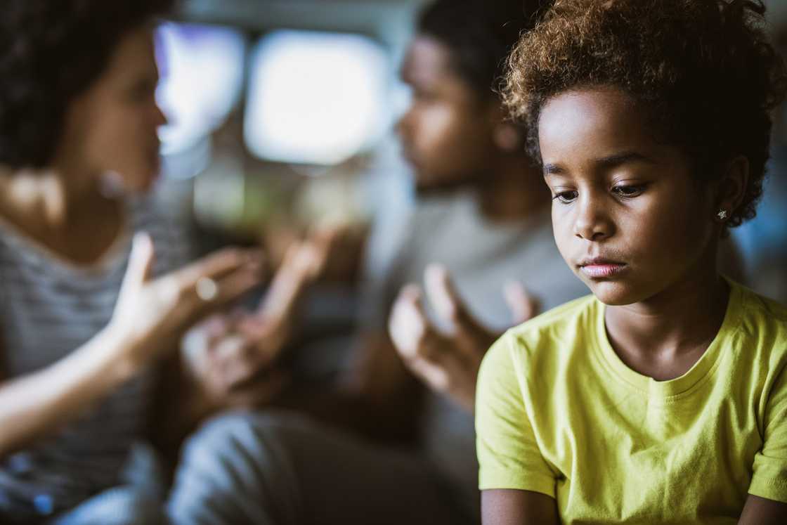 Child in yellow shirt looks down sadly while two adults argue in the blurred background. Child in yellow shirt looks down sadly while two adults argue in the blurred background.