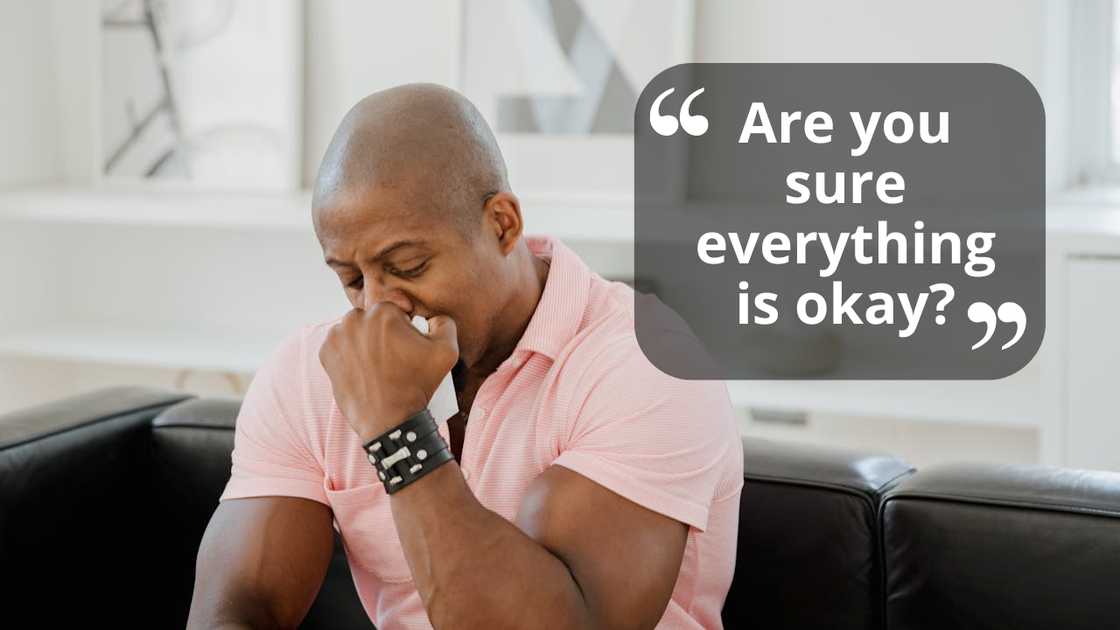 A man sits on a couch with his hand over his mouth, appearing upset.
