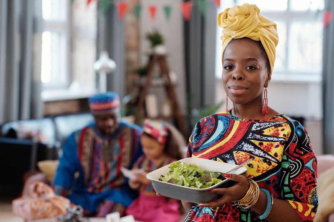 A woman in colourful clothing holds a bowl of salad while her family sits behind her.