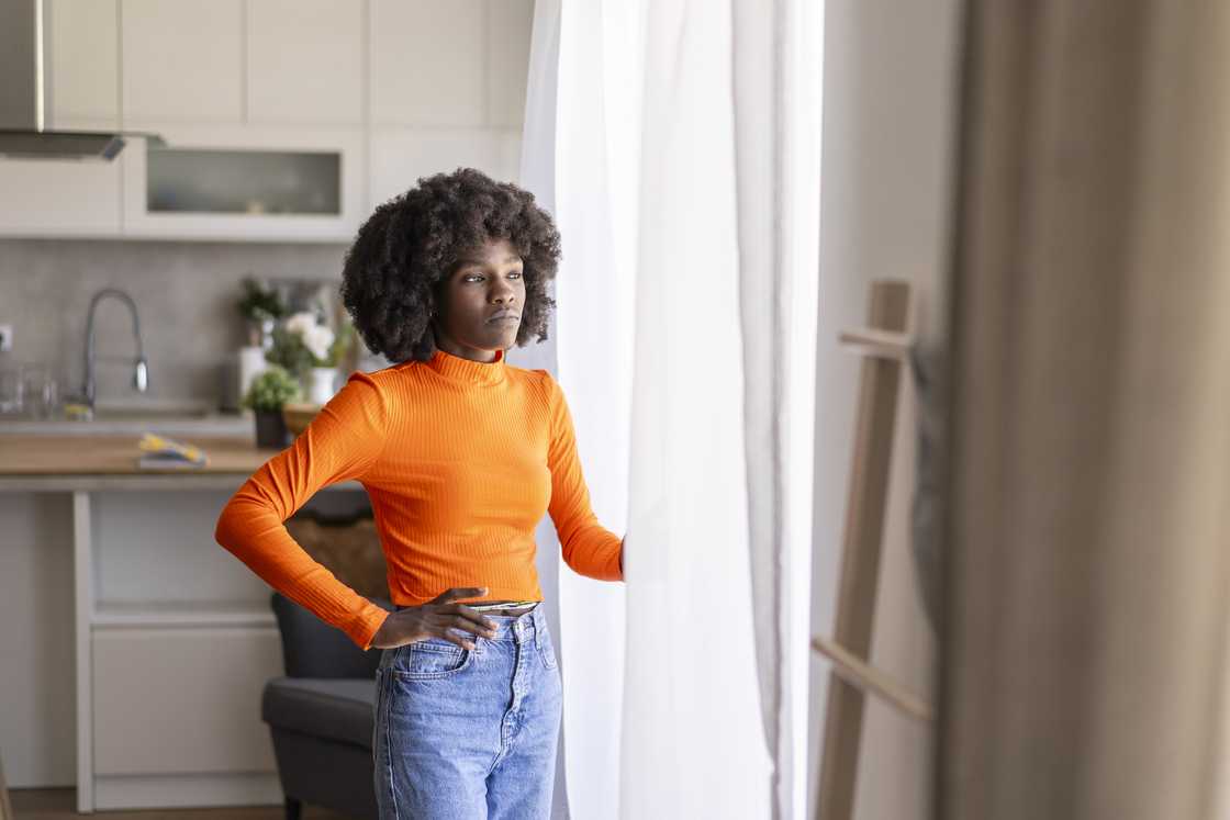 Person in orange crop top stands by a window, holding a sheer curtain. Person in orange crop top stands by a window, holding a sheer curtain.