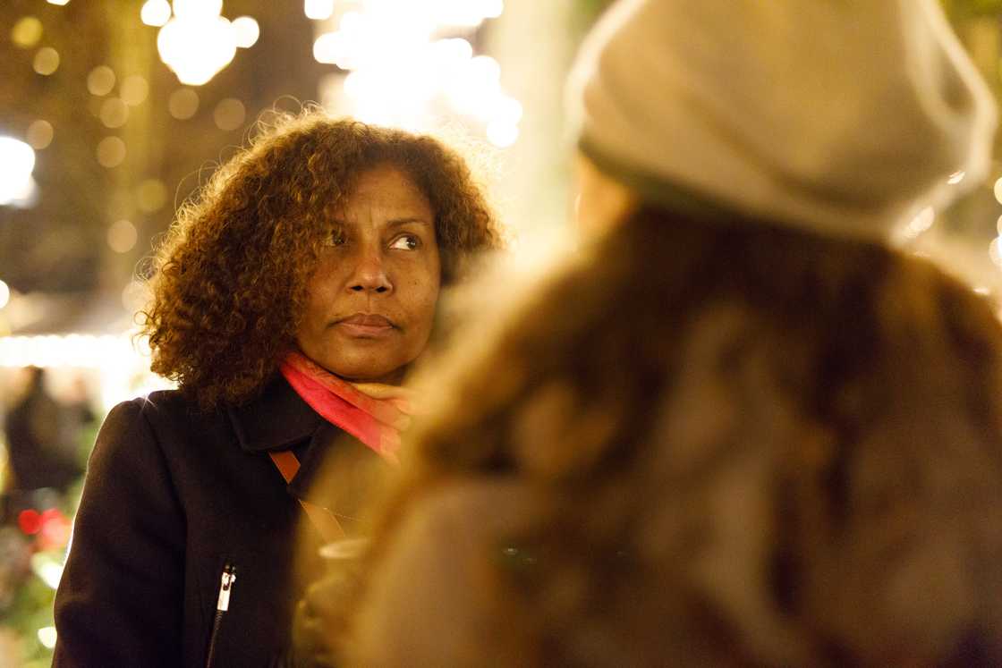 Two people stand outdoors under festive lights, one wearing a beanie and the other a pink scarf.
