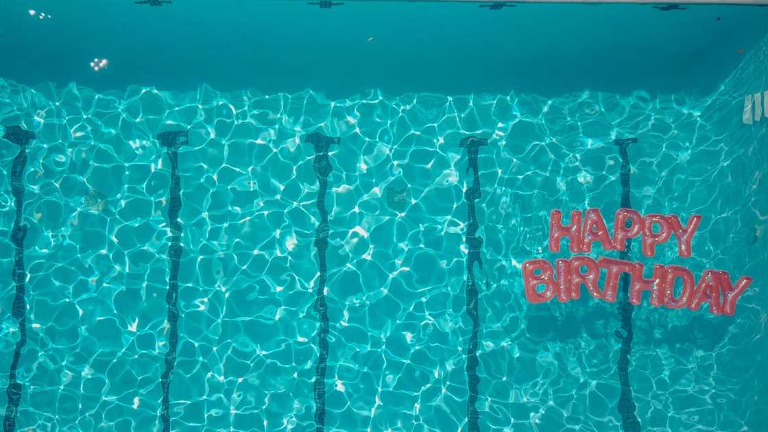 Bright "Happy Birthday" sign bobbing in a pool, with a backdrop of colourful pool floats. Bright "Happy Birthday" sign bobbing in a pool, with a backdrop of colourful pool floats.