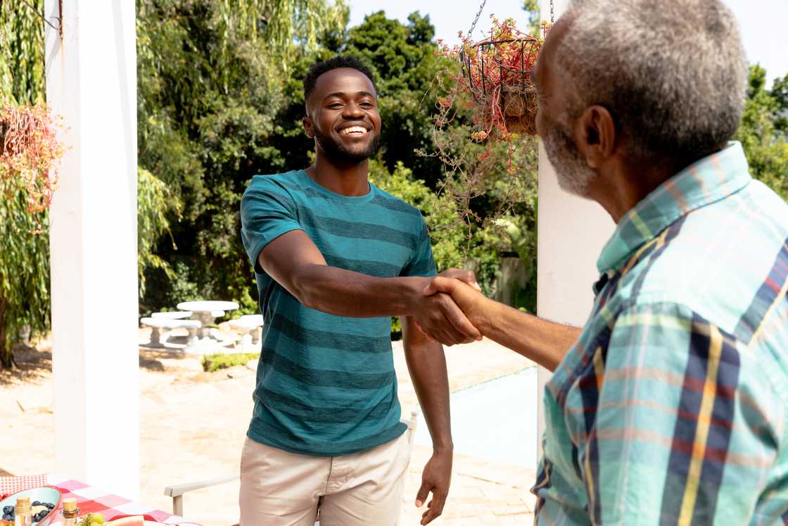 Two people shake hands outdoors near a white column.