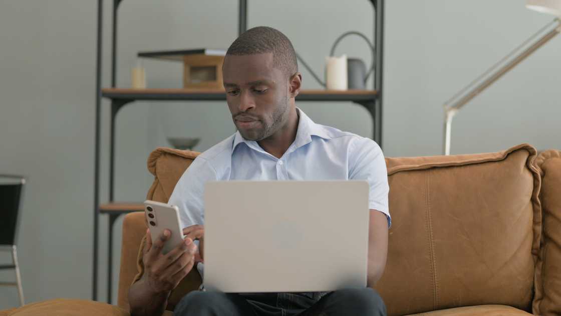 A man using a laptop and smartphone while sitting on a sofa at home