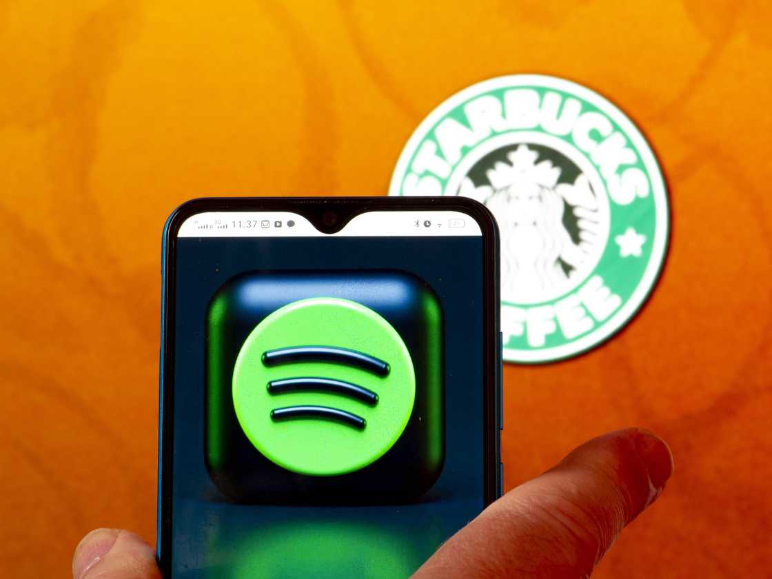 The Spotify logo is seen displayed on a smartphone screen with the Starbucks coffee logo in the background