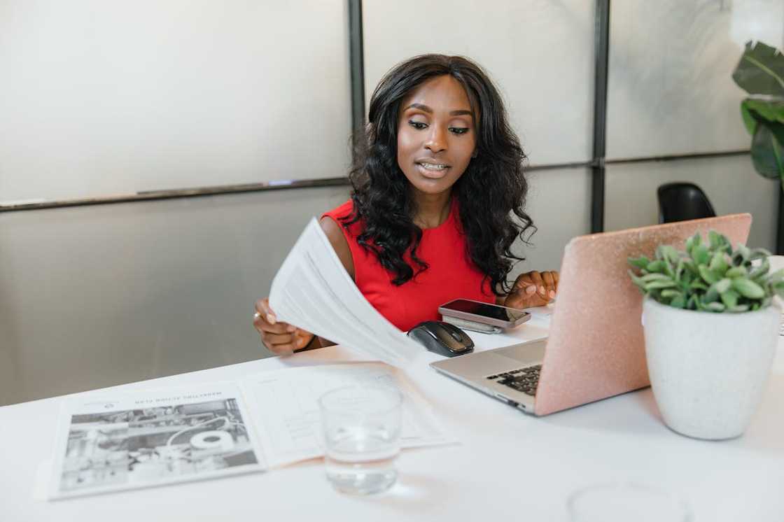 A woman in a red top works at a desk with a laptop and documents. A woman in a red top works at a desk with a laptop and documents.