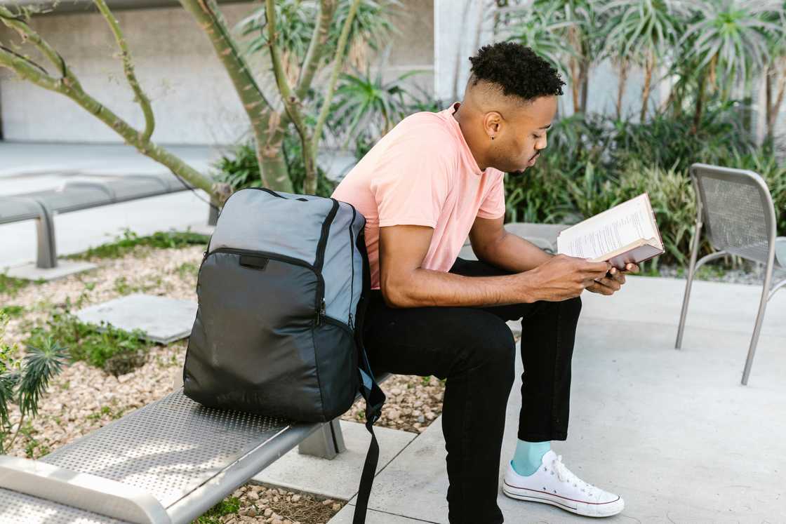 A young man in a pin top and black pants is reading a book on a bench