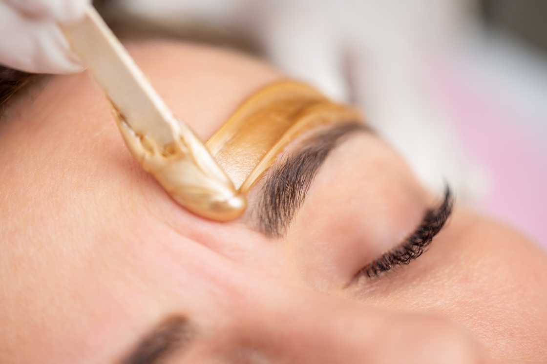 A person applies a gold-coloured wax with a spatula to a woman's face