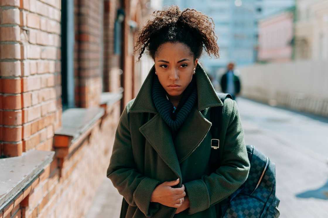 Woman walking outdoors with a serious expression, wearing a coat and carrying a bag.
