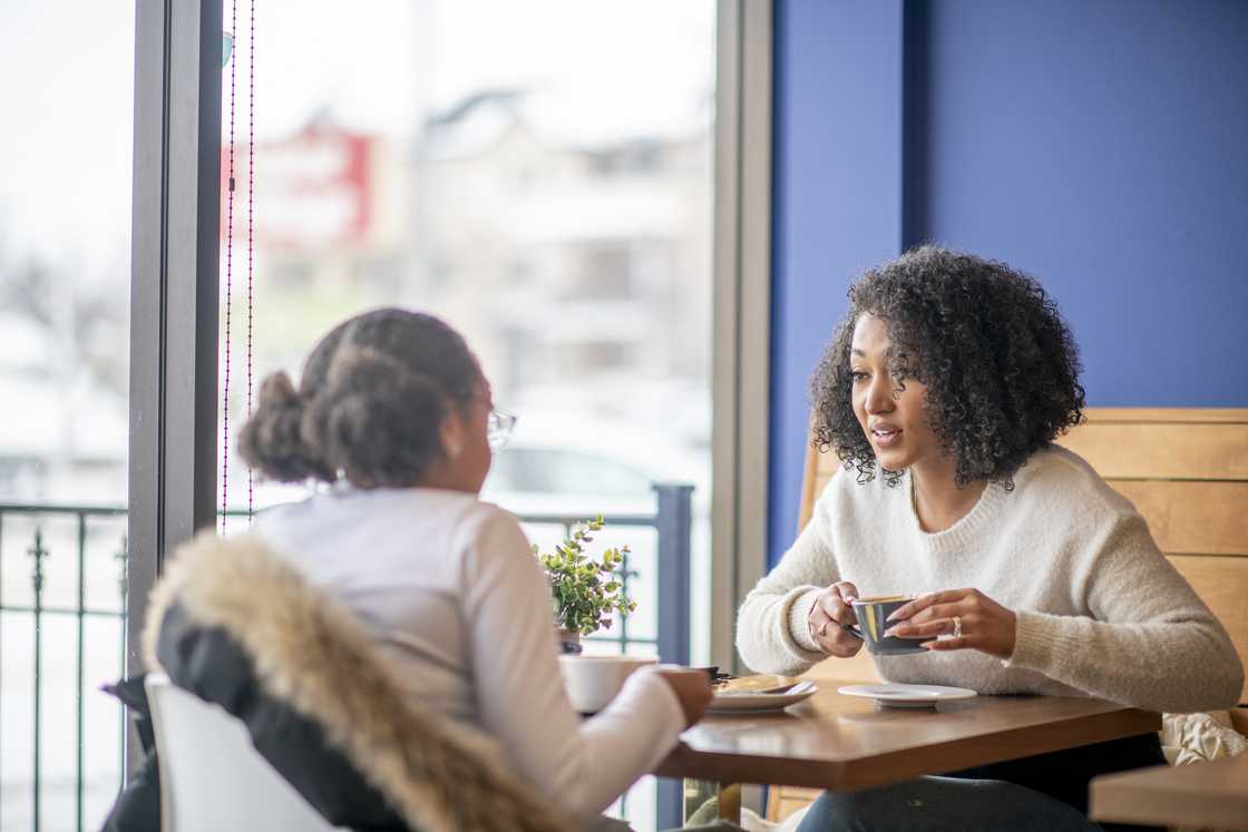 Two people sit at a café table by a window, talking over coffee. Two people sit at a café table by a window, talking over coffee.