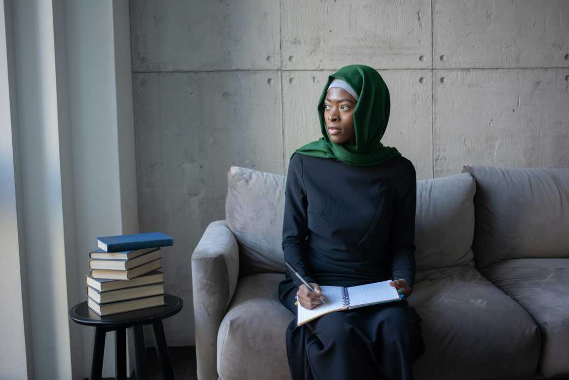 A Muslim woman in a hijab sitting on a grey couch by a window writing on a notebook