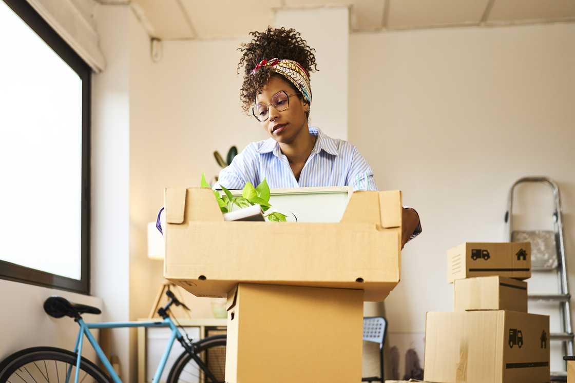 A woman standing in the living room of an apartment, carrying a moving box