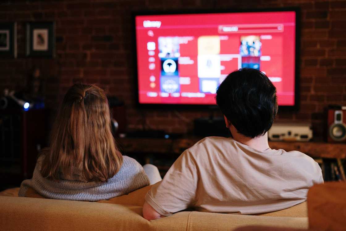 A couple is seated on a brown coach watching television. A couple is seated on a brown coach watching television.