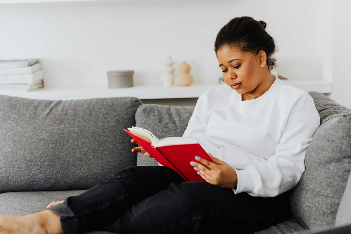 A woman in a white top and black pants is reading a book on a grey sofa