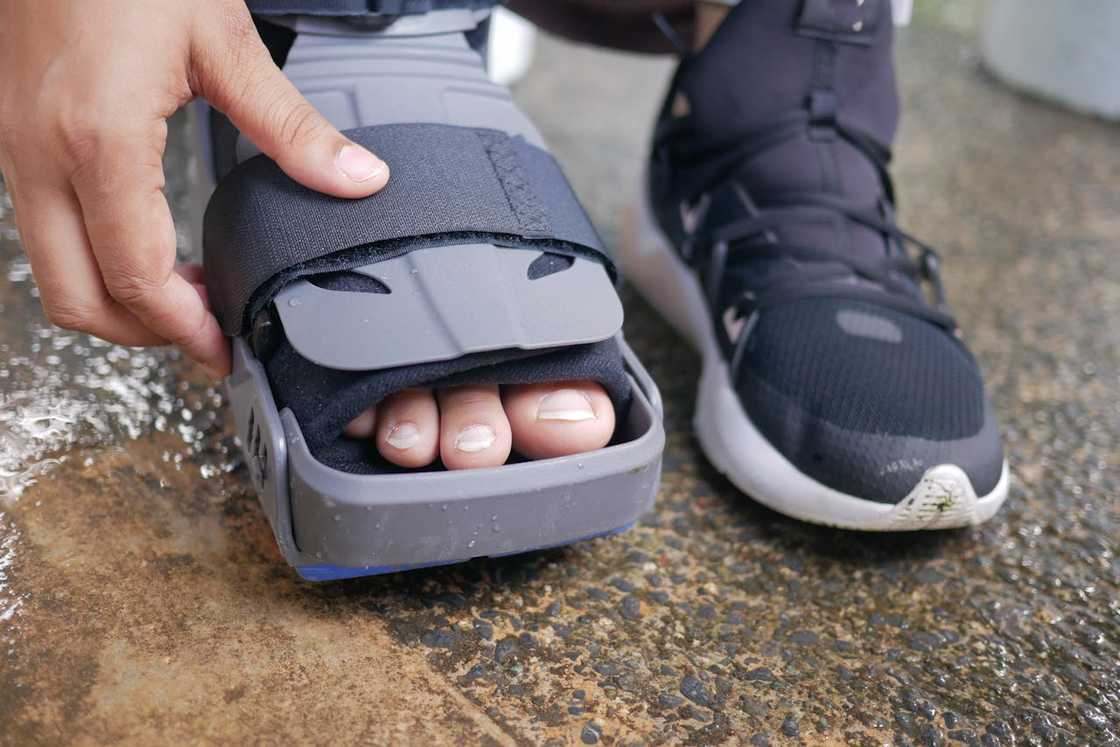 A person adjusts a medical walking boot on one foot while wearing a trainer on the other.