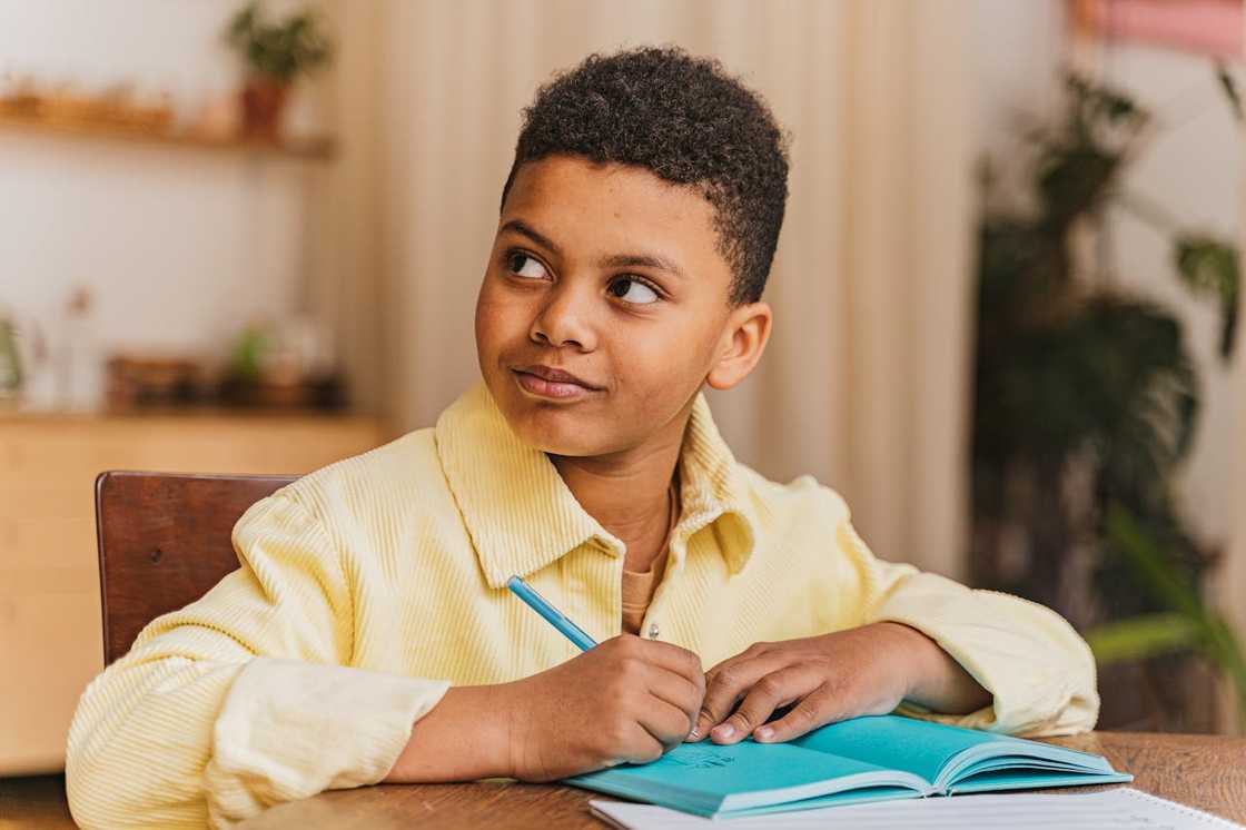 A young boy writes in a blue notebook while looking off to the side thoughtfully.